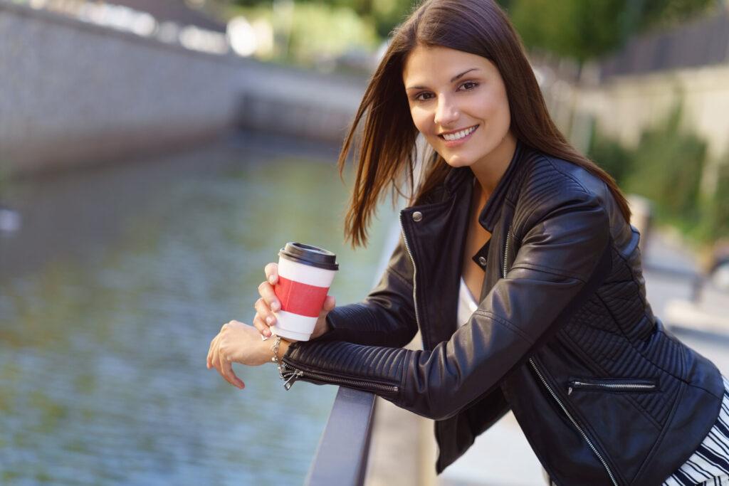 Frau mit einem Becher Kaffee in der Hand lehnt sich entspannt an ein Geländer in der Stadt