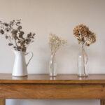 Nahaufnahme künstlicher Blumen und Blätter in Glasflaschen und weißer Kanne auf Beistelltisch, aus Eichenholz vor beiger Wand.