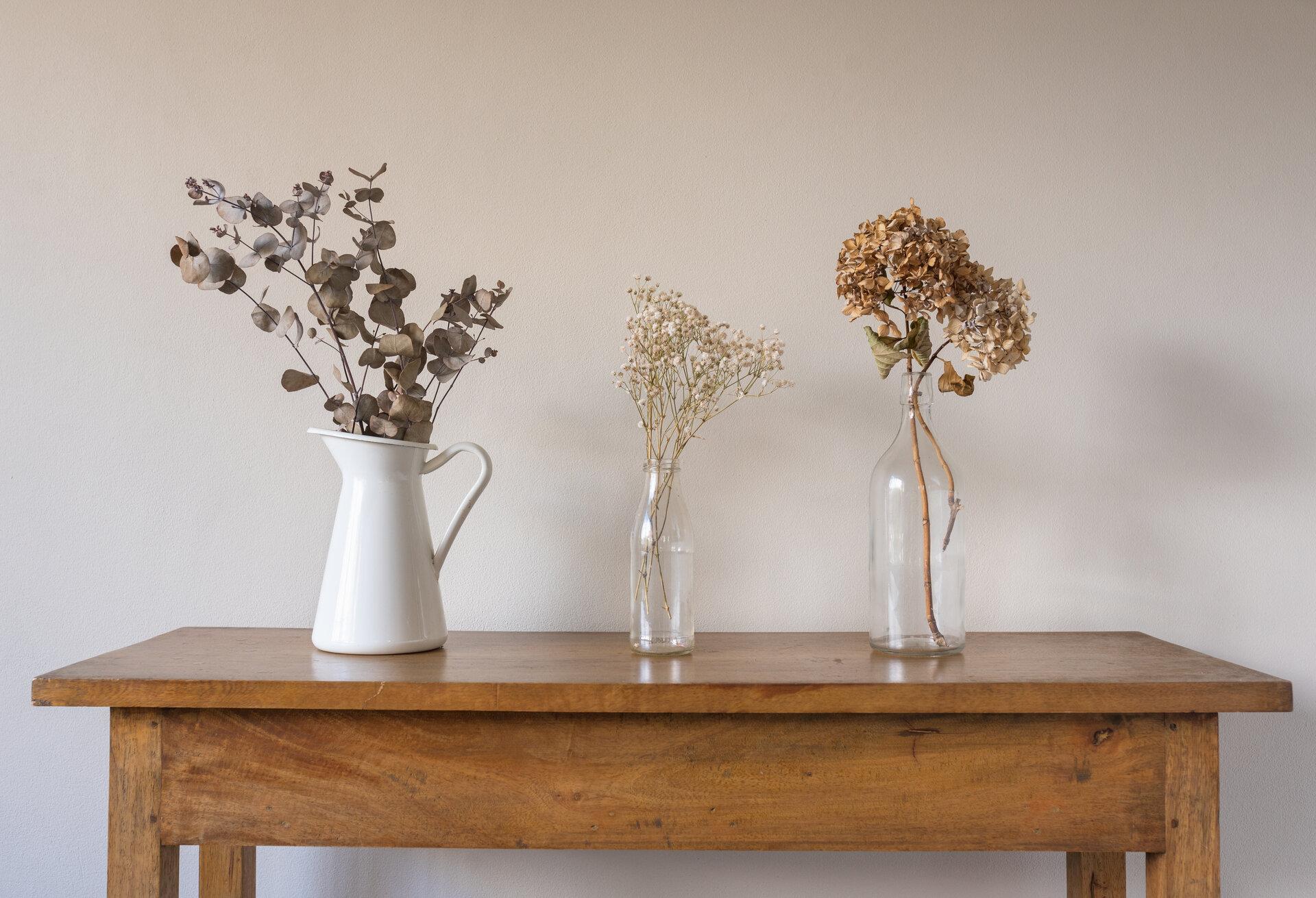 Nahaufnahme künstlicher Blumen und Blätter in Glasflaschen und weißer Kanne auf Beistelltisch, aus Eichenholz vor beiger Wand.