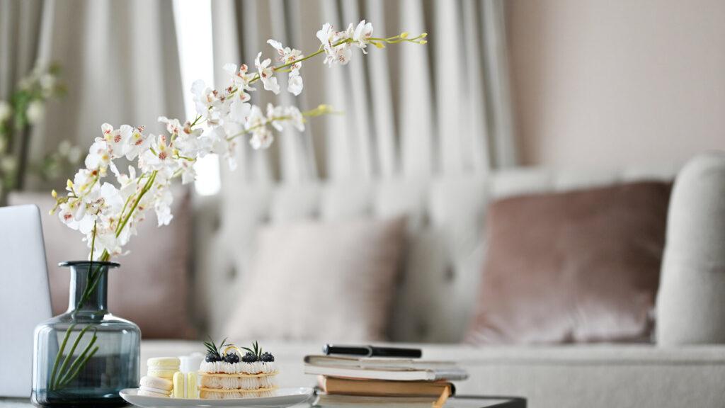 Modernes Wohnzimmer mit bequemem Sofa, künstlichen Blumen in einer Keramikvase, Buch, einem Dessertteller und Dekor auf dem Couchtisch.