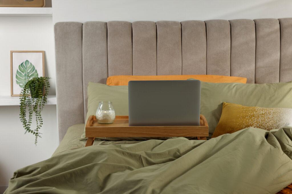 Praktisches Holztablett mit modernem Laptop und brennender Kerze darauf, steht stabil auf dem Bett im Schlafzimmer.