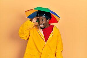 Afrikanisch-amerikanischer Mann mit gelbem Regenmantel und Regenbogenmütze, sehr glücklich und lächelnd, hält sich die Hand an den Kopf und hält suchend Ausschau.