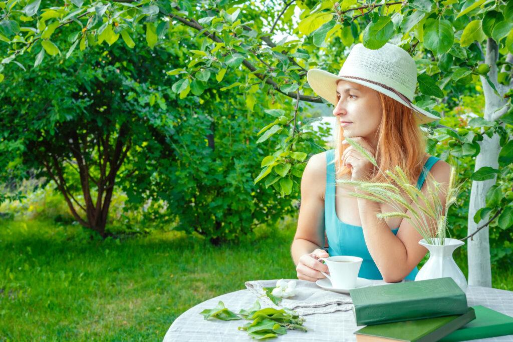 Frau in einem Kleid, mit Strohhut und mit einer Tasse Tee in der Hand, sitzt am Tisch im Garten an einem Sommermorgen.