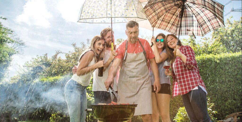 Menschen grillen im Garten bei Sonne und Regen unter Regenschirmen mit Doppelstegplatten-Schutz.